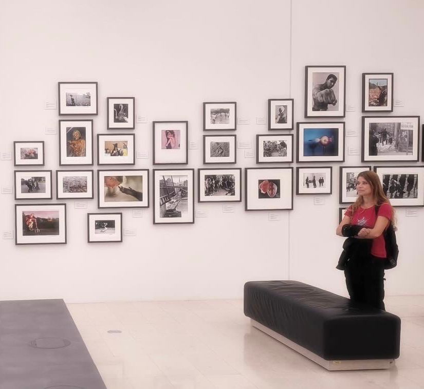 In een grote zaal, van een afstand gefotografeerd. Ik sta en kijk naar een groot scherm, achter mij een witte muur met verschillende iconische foto’s van grote gebeurtenissen in de geschiedenis, zoals een foto van Einstein, een foto van Jane Goodall met de hand van een aap, een foto van Ghandi etc. op A4-formaat.