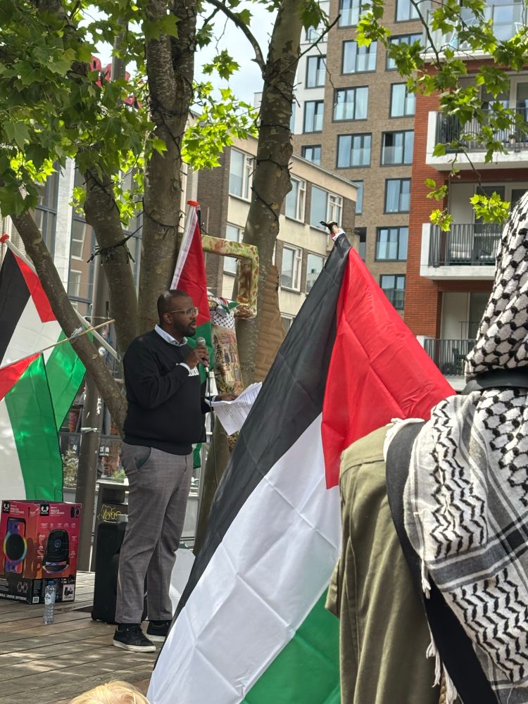 Foto van raadslid Abdulahi Hussen die een zwarte trui draagt en grijze broek en een microfoon in zijn handen heeft. Hij staat op Plein ‘44 in Nijmegen. Ik fotografeer hem vanaf de zijkant. Voor mij staan mensen met de vlag van Palestina en de kenmerkende wit-zwarte keffiyeh.