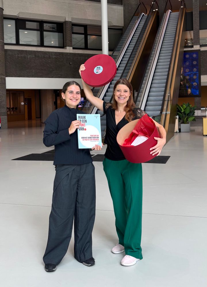 Foto in de Staten-Passage van de Tweede Kamer, voor de roltrap. Ik sta daar met een afgevaardigde van Women Inc, zij (donkergrijze vroeg en donkere trui) links en ik (zwart shirt en groene broek) rechts. Ik heb een ronde en rood gespoten hoedendoos vast. Daarin papieren met de boodschap.