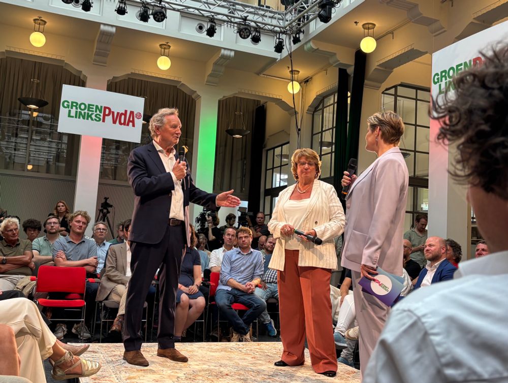 Foto van de zaal in het Nutshuis in Den Haag. Op het podium staan Bram van Ojik en Mariette Hamer, de voorzitters van de programmacommissie, met Mariëlle Vavier die de bijeenkomst modereerde. Het podium staat in het midden van de zaal, met aan alle kanten mensen eromheen, in een vierkant. Zij zitten op rode stoelen. Ik neem de foto en zit schuin achter Jesse Klaver van wie op de foto het achterhoofd te zien is.