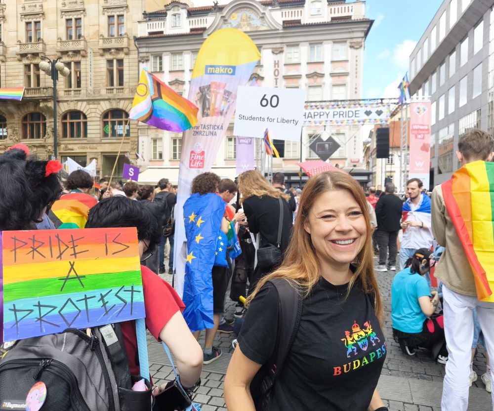 Foto van mijn in Praag, tussen mensen met regenboogvlaggen en andere vrolijke kleding. Ik draag mijn zwarte shirt waar in regenboogkleuren Budapest op staat. Links van me (op de rug gefotografeerd) iemand met een bord waarop staat “Pride is a Protest”. Voor ons een podium met een grote banner met de tekst “Happy Pride”