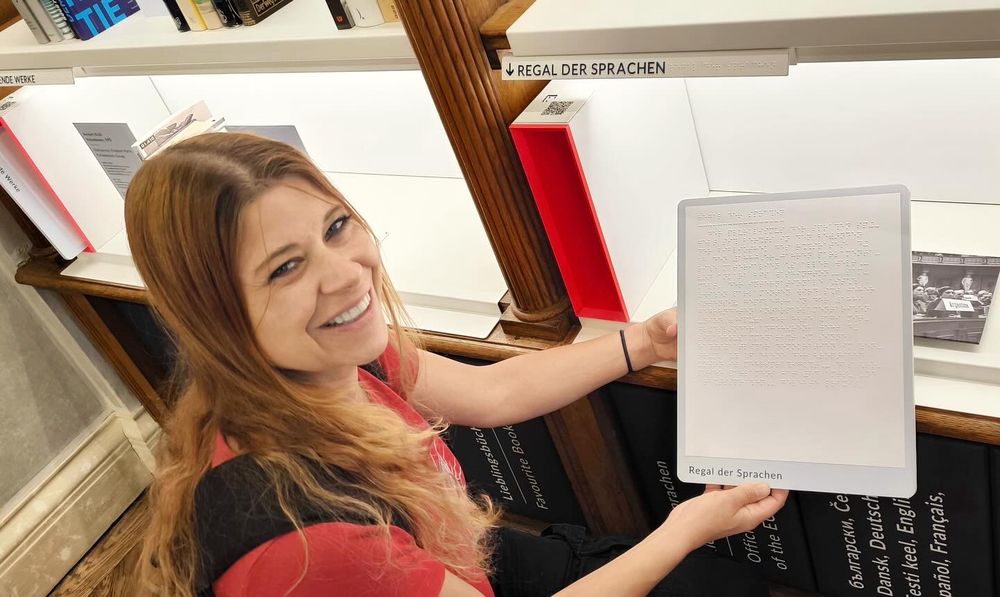 Foto van mij in de bibliotheek van het parlement van Oostenrijk. Op de achtergrond informatieposters (onleesbaar op de foto). Ik heb mijn haar los, heb een rood shirt aan en rugzak en zit gehurkt. In mijn hand een kunststof plakkaat op a4-formaat met braille opdruk.