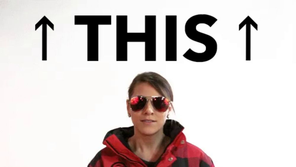 a woman wearing sunglasses and a red jacket is pointing up with the words " this " above her