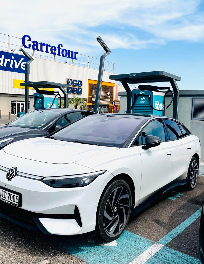 A white electric Volkswagen car is parked next to a charging station in front of a Carrefour store. The scene includes clear skies and a visible sign for the Carrefour location.