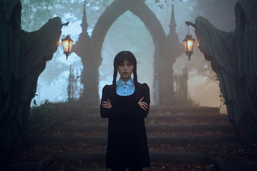 A girl wearing black and braids with her arms crossed standing on a stairway with statues that look like hooded humans on both sides that are holding lanterns  