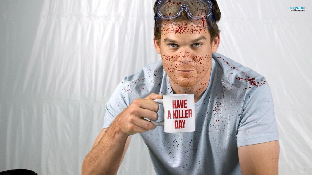 A man in a light blue shirt, wearing goggles on his head with red splatter on himself while holding a cup of coffee that says “Have a killer day”