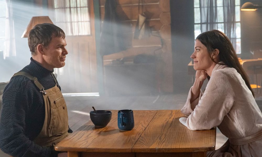 Man in overalls sitting at a table across from a woman at a dinning table in a cabin.