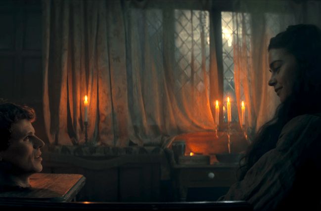 A woman sitting in a candle lid room staring at the human head that is set on a table across from her