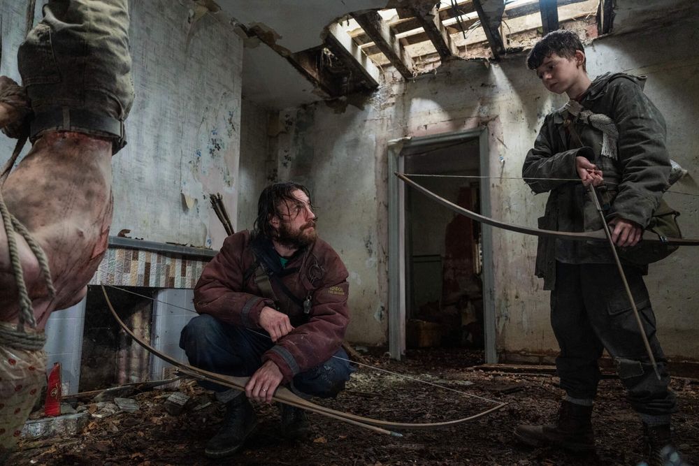 A man and a kid with bows and arrows standing in an abandoned building.  
A body is hanging upside down on the left side of the room.  