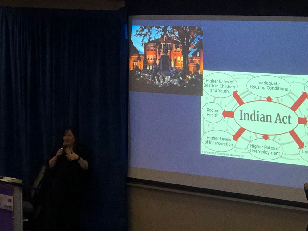 Carolyn Roberts speaking in front of a projected screen showing a slide with a diagram with Indian Act in its centre arrowing out to many implications