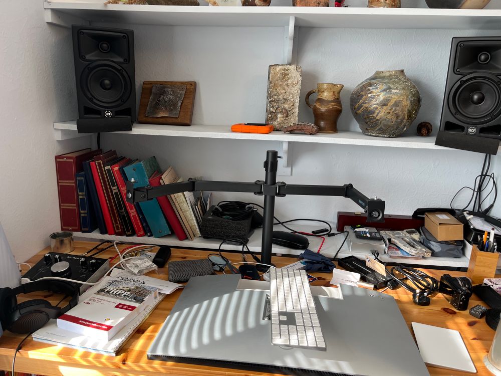 A half-dismantled workstation, with empty screen stands still reaching out above a desk, with one computer monitor facedown on the desk below it. Various books and albums are visible, including a PHPP manual to the left of the desk. Pots and speakers are on the shelves behind.