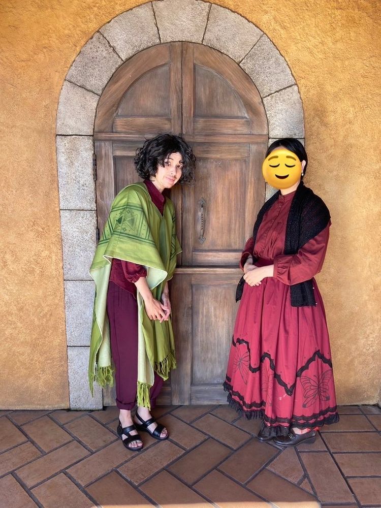 ディズニーシー、昼間。ポンデ・ヴェッキオの端寄り。黄土色の壁に、石組みの枠のついた古い木製の扉がある場所を正面から捉えた構図。扉の前に、ブルーノ仮装の帆川とアルマ仮装のとと氏が間に人ひとり分ほどの距離を置いて立っている。ブルーノは少し背を丸め、体の前の下の方で軽く指を組み、眉を下げて弱い笑み。アルマは姿勢よく立ち、腰の前で品よく手を重ね合わせている（顔は穏やかに目を伏せた表情のスタンプで隠されている）