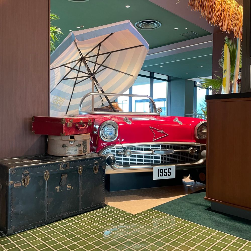 「1955 東京ベイ by 星野リゾート」のエントランス（このポストの他の写真も同じ場所）
落ち着いたグリーンのタイル床の上、柱とフロントカウンターの間に挟まれるようにして、鮮やかな赤色のレトロなオープンカーが設置されている。ナンバープレートは「1955」車にはビーチで使う大きなパラソル（白地に水色の縞模様）が広げてさされている。周囲には年季の入った大きなトランクや衣装ケース、サーフボード、南国らしい木の装飾。