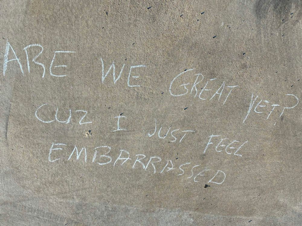 Chalk writing on the sidewalk saying “Are we great yet? Cuz I just feel embarrassed.”