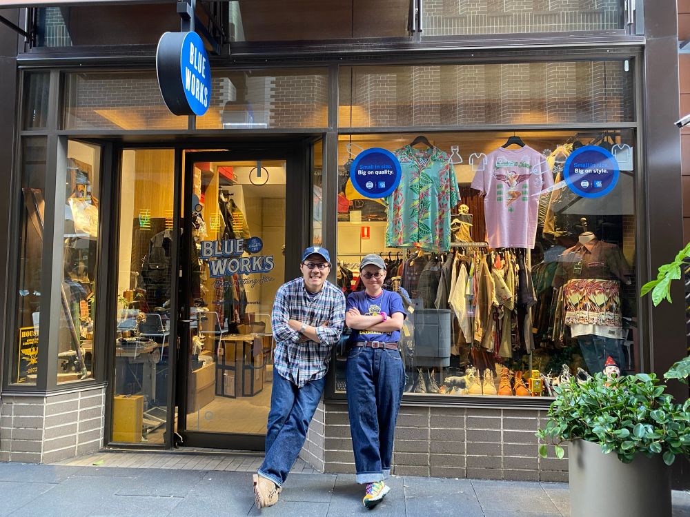 Dang and Steve stand casually outside a shop called “BLUEWORKS.” Both are wearing caps, denim jeans, and relaxed outfits. Steve leans against the doorframe with arms folded, wearing a plaid shirt and beige shoes. Dang stands with arms crossed, wearing a navy graphic T-shirt, dark loose-fit jeans, colourful sneakers, and round sunglasses. Behind them, the shop window displays patterned shirts, clothing racks, and accessories. The scene has a friendly, easygoing street-style vibe.