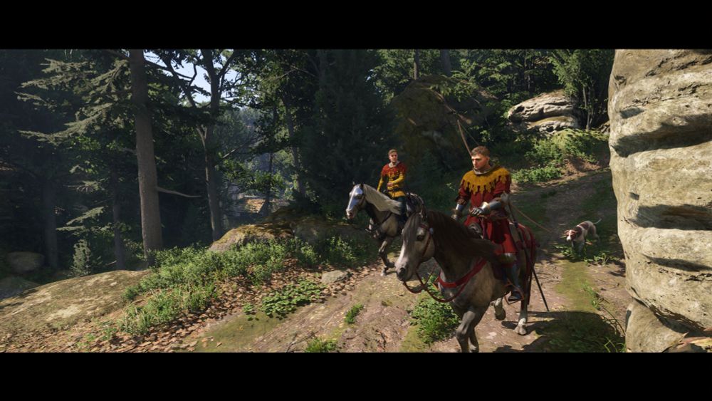 screenshot of kingdom come deliverance: II showing Hans and Henry riding in a forest. Hans is wearing his signature yellow pourpoint and red hood, Henry is wearing a red top and a yellow hood. 