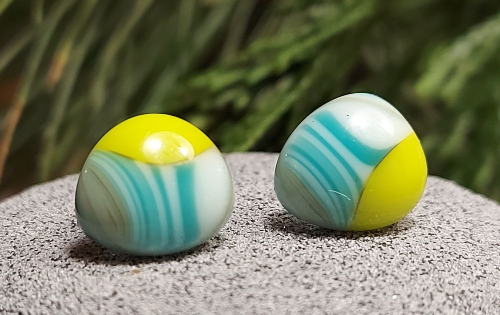 Pair of stud earrings which are in wonky, i.e. not round but in the shape of very rounded triangles. They are made of non transparent or opalescent glass. One tip of the triangle is lime green glass, and the rest is striped glass going from grey to white and light blue stripes to white. So second tip is greyish, the third tip is white and the middle is striped white and light blue glass.
Photographed on rough grey surface with greenery in the background.