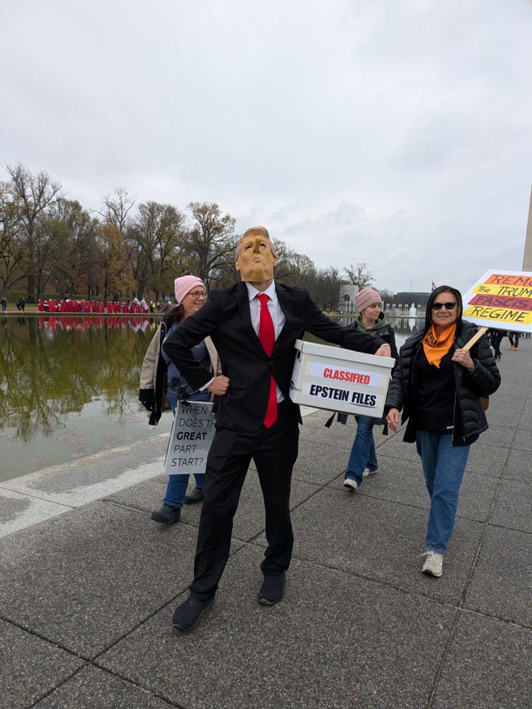 A group of people walking one is dressed in a suit and donOld mask carrying a file box labeled "Epstein files"