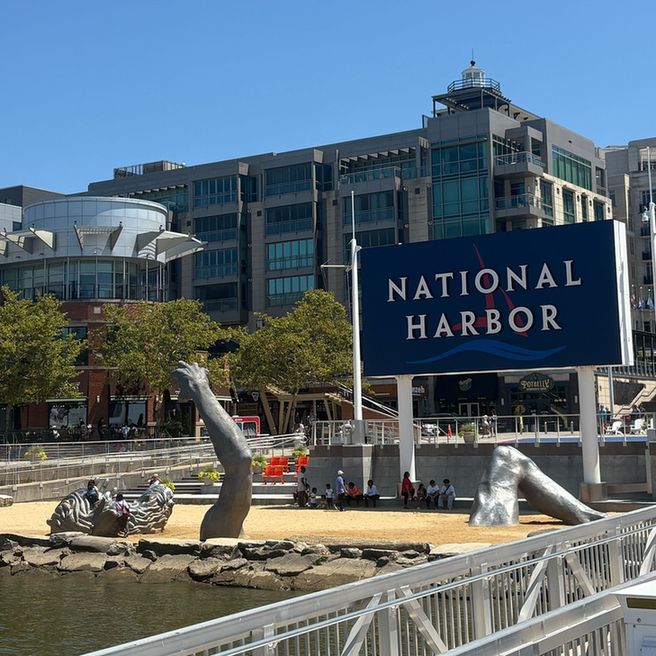 A view of the National Harbor with a large sign reading 'NATIONAL HARBOR' in white letters on a blue background. In front of the sign, a sandy area features parts of a large silver sculpture emerging from the ground, including an arm and a head. Modern buildings and trees line the background under a clear blue sky.