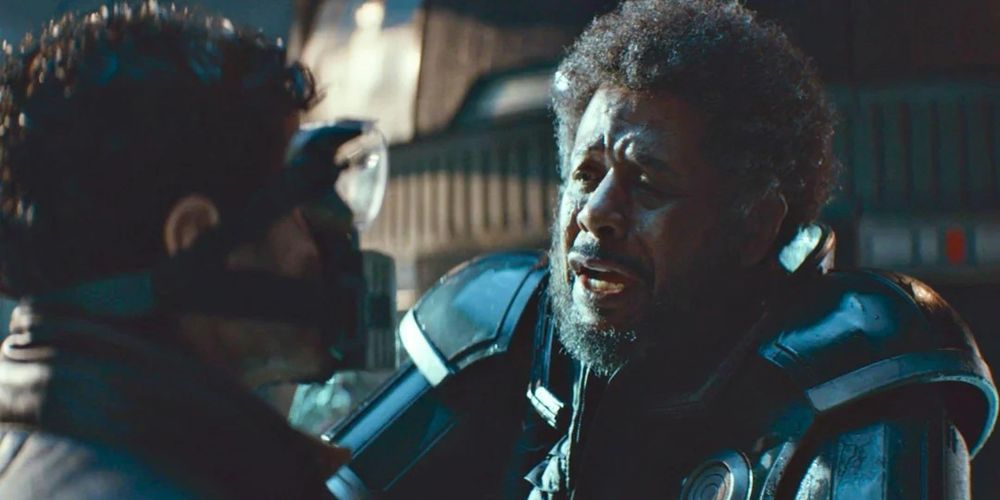 Saw Gerrera confronts Willmon in Andor, gripping him by the shoulders in a tense moment. Willmon wears a breathing mask while Saw, clad in his distinctive armored suit, looks distressed and intense amid a dim, industrial setting.