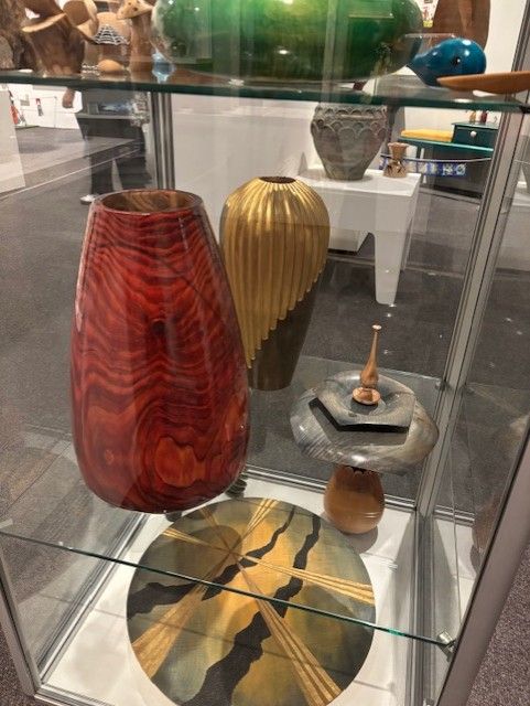 Display case containing lacquered and gilded wooden objects: a lacquered green bowl, a lacquered rust-red vase, a vase with partial gilding in vertical ridges, a grey item that looks a bit like a sundial, a large flat circular item with a gold and black design that looks a little like sun rays and clouds. 