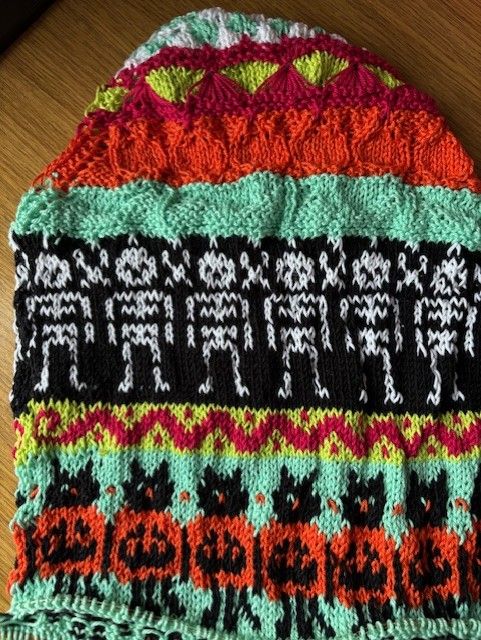 The chart from previous days, now with a long colourwork section. The background is mint green, there are black cats with green eyes and orange noses, standing and holding pumpkins as large as their bodies. 