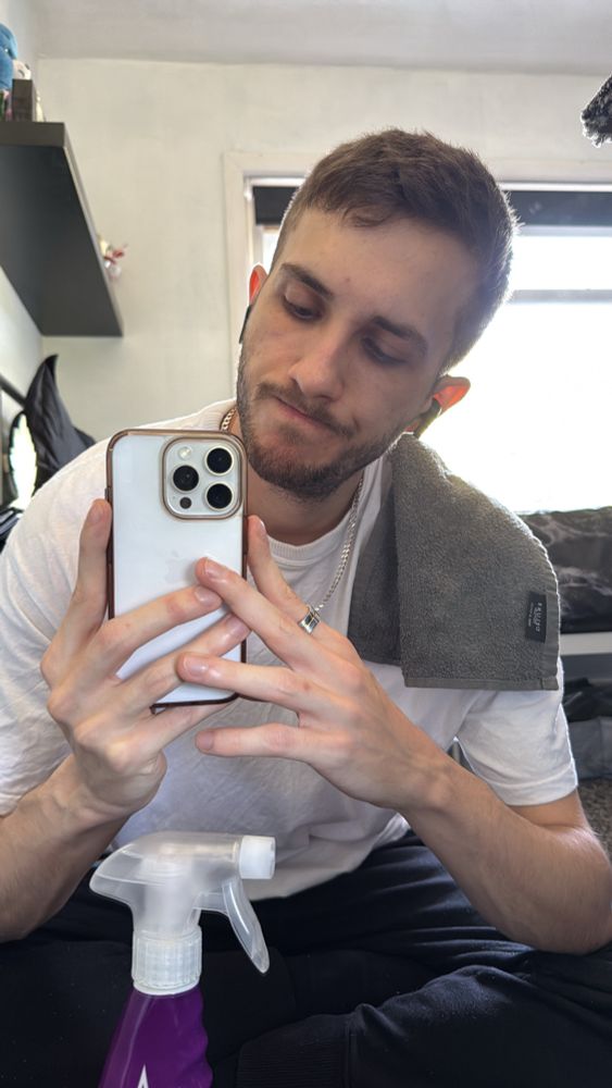 A man sat cross legged in front of a mirror with a cloth over his shoulder and a mirror/glass spray bottle.