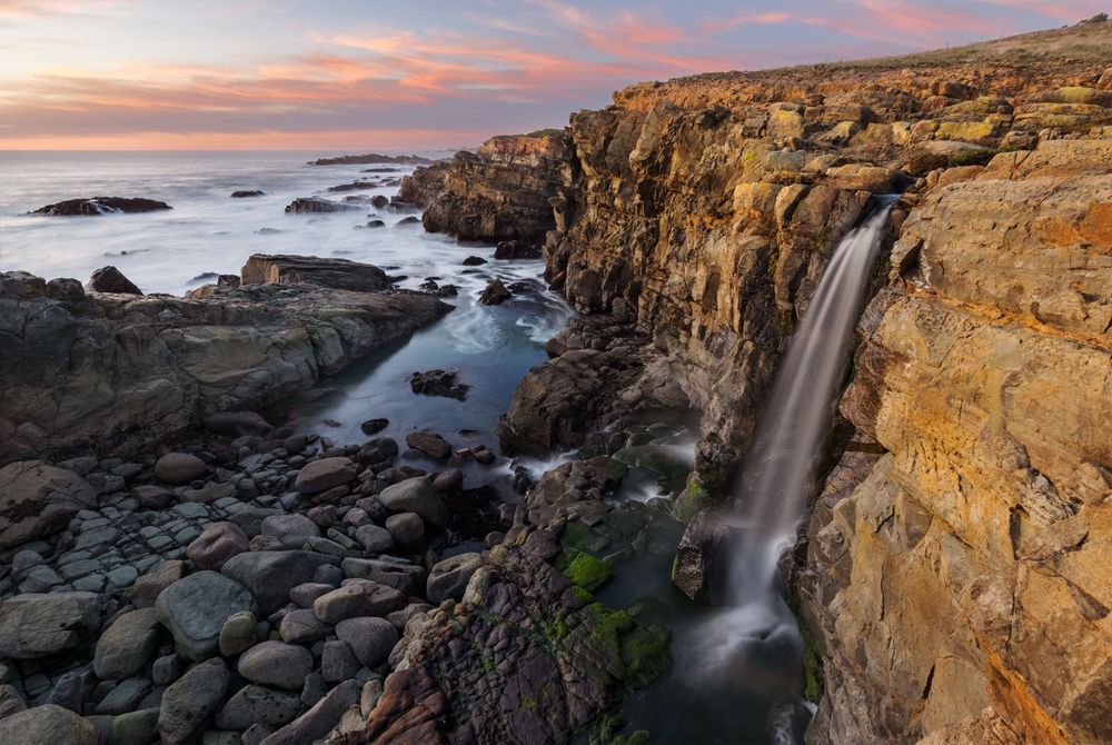 Long exposure of a rocky coastline at sunset. From the cliff on the right side, a waterfall drops down onto rocks below and flows into the ocean in the background. Boulders lie on the bottom left and colorful clouds are in the distance at the top of the scene