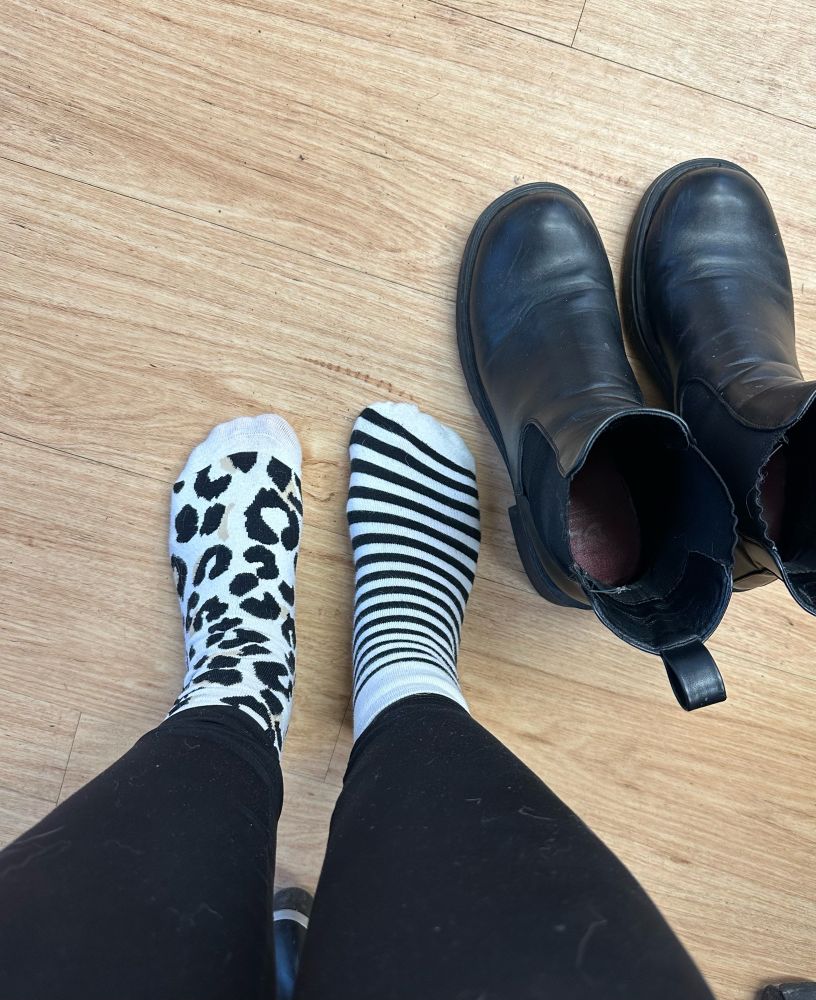 Feet wearing mismatched socks, one striped and one leopard print, on a hardwood floor. Pair of black boots on the floor beside.