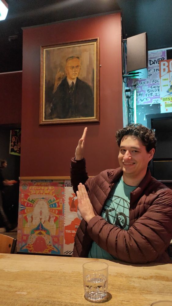 A smiling man pretends to hold something up. He is in front of a portrait.