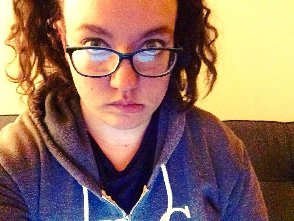 Selfie of Christine biting their lip and looking at the camera. They're wearing a hoodie and glasses and their dark brown curly hair sits at their shoulders. They look their age, which is 25.