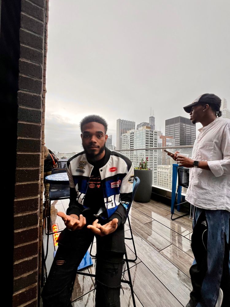 goat sitting on a patio wearing a blue, black and white biker jacket a black t shirt and black pants. You can see some of Chicagos skyline behind him and there’s another person in the picture not facing the camera wearing a black and white hat a white shirt and denim pants 