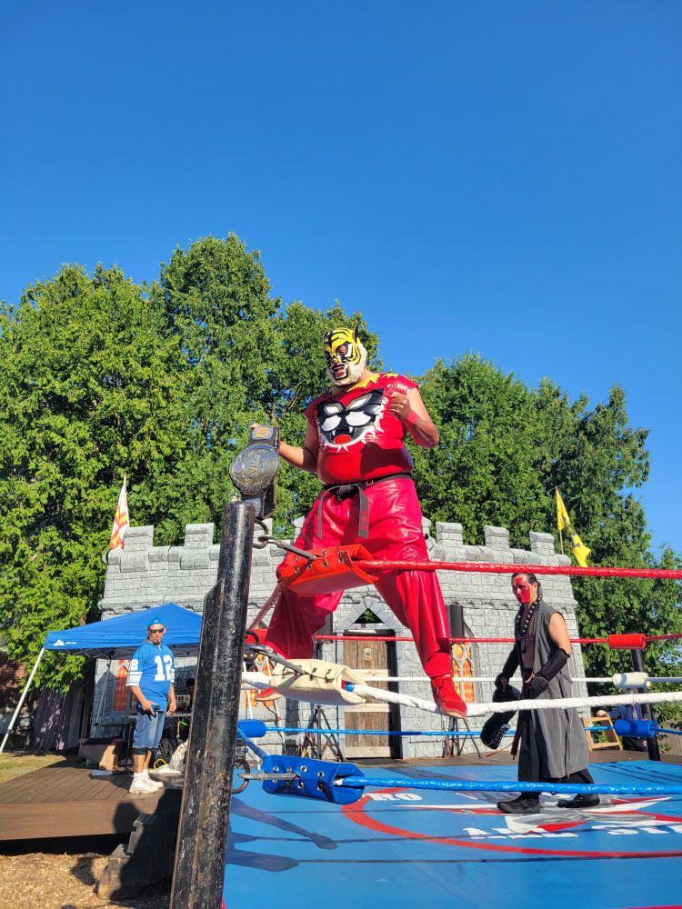 A wrestler in a red outfit with a tiger mask standing on the ropes of a wrestling ring, holding a championship belt