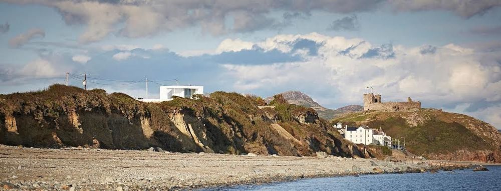 a panoramic image shows a modernist home built on top of, and partly concealed by, this big brown sloping cliff toward the sea