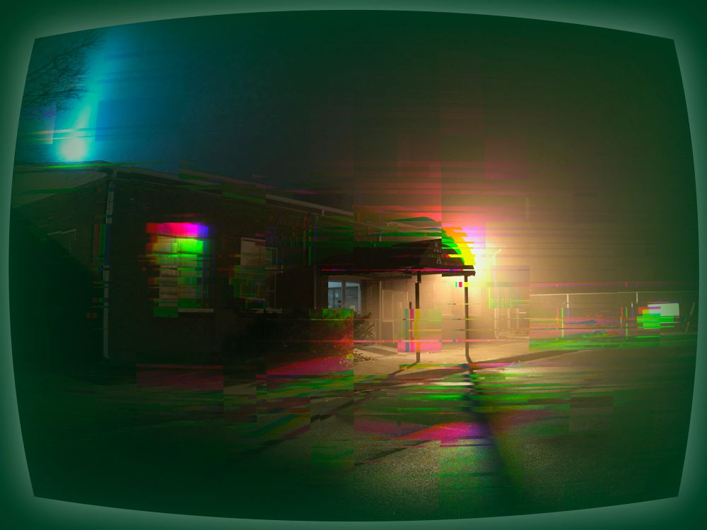 A photo of the original terminal building at the Raleigh County Memorial Airport (now a suite of offices), taken at night and suffused in an eerie green glow, as well as digitally glitched to obscure or scramble some of the building details and signage. One of the more prominent glitch effects is the splitting of the few bright light sources into prismatic strips; this effect is concentrated around one window of the building and the backlit edges of an awning over the entryway—held up by two thin metal poles. In the foreground, the surface of an asphalt parking lot gleams with puddles of moisture lit by the same bright light behind the awning, and these muted light effects too are split into dimmer jagged prisms of color. A chain link fence behind the building and some features of the signage and entryway are glitched horizontally into distorted shapes, and a laser-bright beam of concentrated blue light is beaming upward or lancing downward in the upper left corner of the frame, appearing to strike (or emanate from) the roof of the building. The entire dark scene—suffused in the eerie green glow and punctuated by the blue beam and few prismatic color- and structure-glitches—is warped into the shape of an old CRT TV screen, and framed with a glowing dark green border.