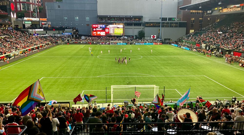 A view of Providence Park during a Portland Thorns soccer game, taken from section 107. Queer and trans pride flags wave in the foreground.