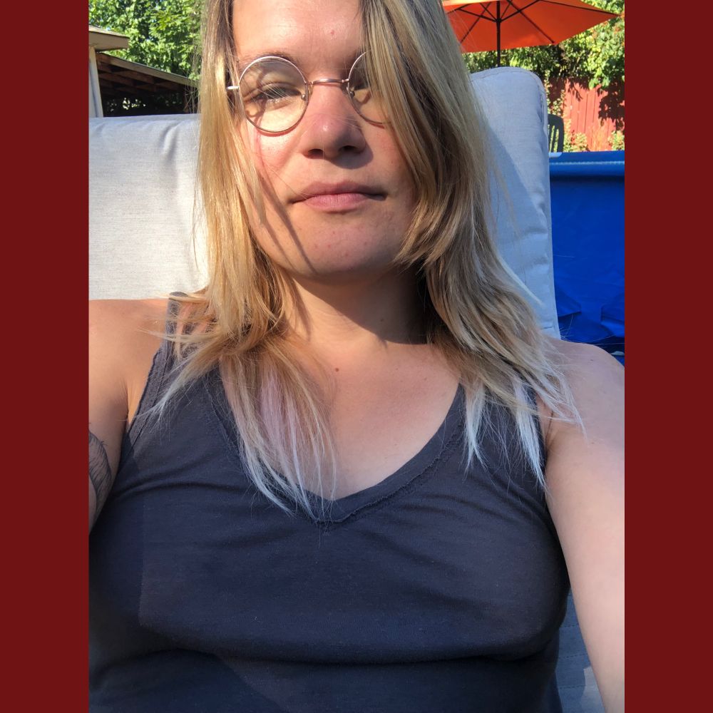 selfie of a white trans woman looking at the camera with sun in her eyes. she’s got long lightened hair and a dark grey dress on