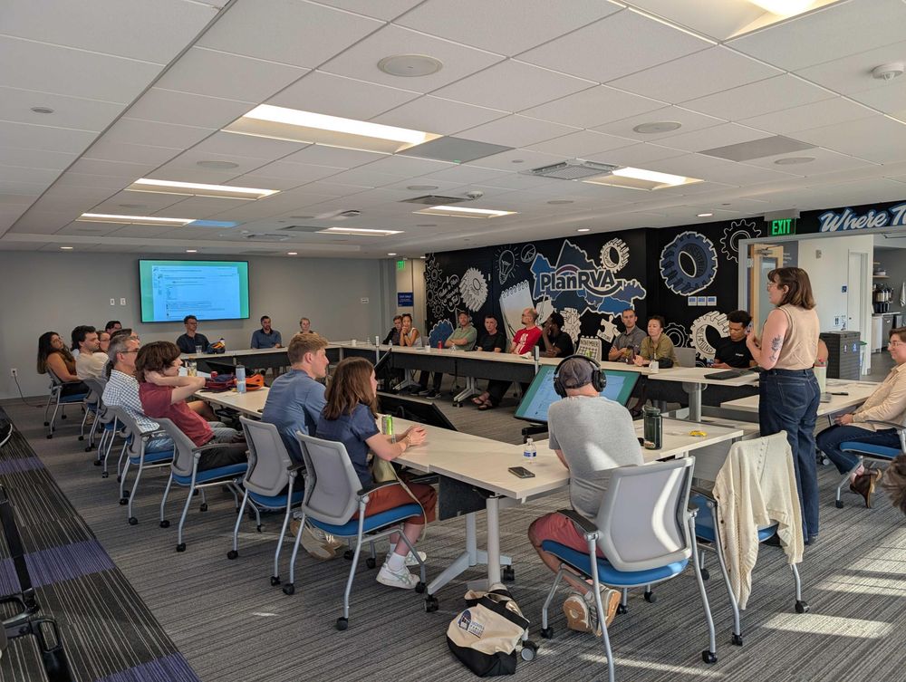 A female speaker addresses an audience seated in a circle around a room. On the opposite side of the room, a TV on the wall shows the speaker's current slide.