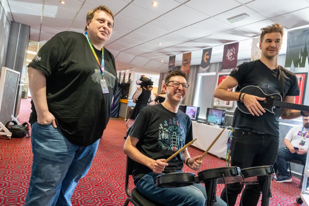 Mike Chapman sitting on a Rockband Drumkit, playing together with Loki the Sea of Thieves Monkey Island Parkour only by using the guitar and drumkit to control the character. Leator is standing next to them.