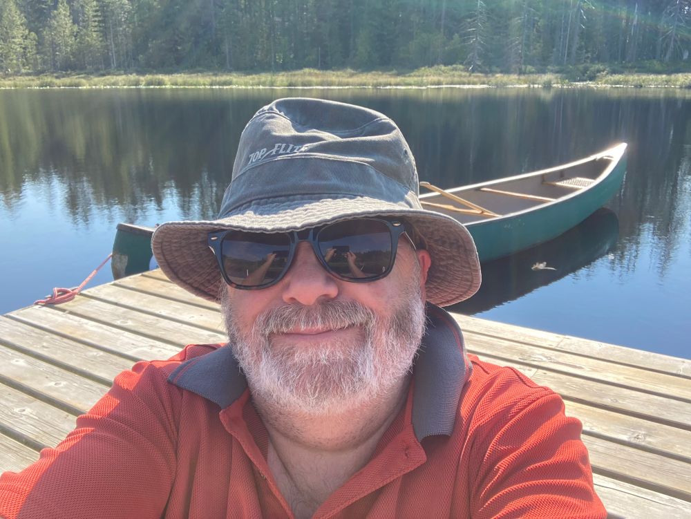 Sean sitting on the dock in a floppy hat and sunglasses with “Battlecanoe Potemkin” tied off in the background