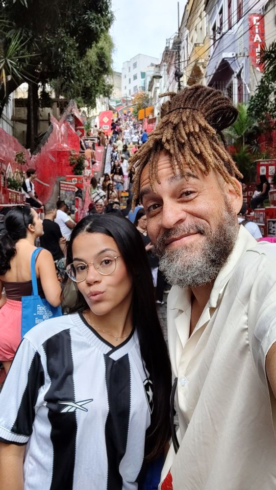 Selfie eu e minha filha. Ela pele parda, cabelos pretos lisos e longos, óculos de grau e camisa do Botafogo. Eu pele parda, cabelos com dreads presos pro alto e caindo pela testa, camisa de botão bege clarinha. Atrás uma multidão na Escadaria Selaron.