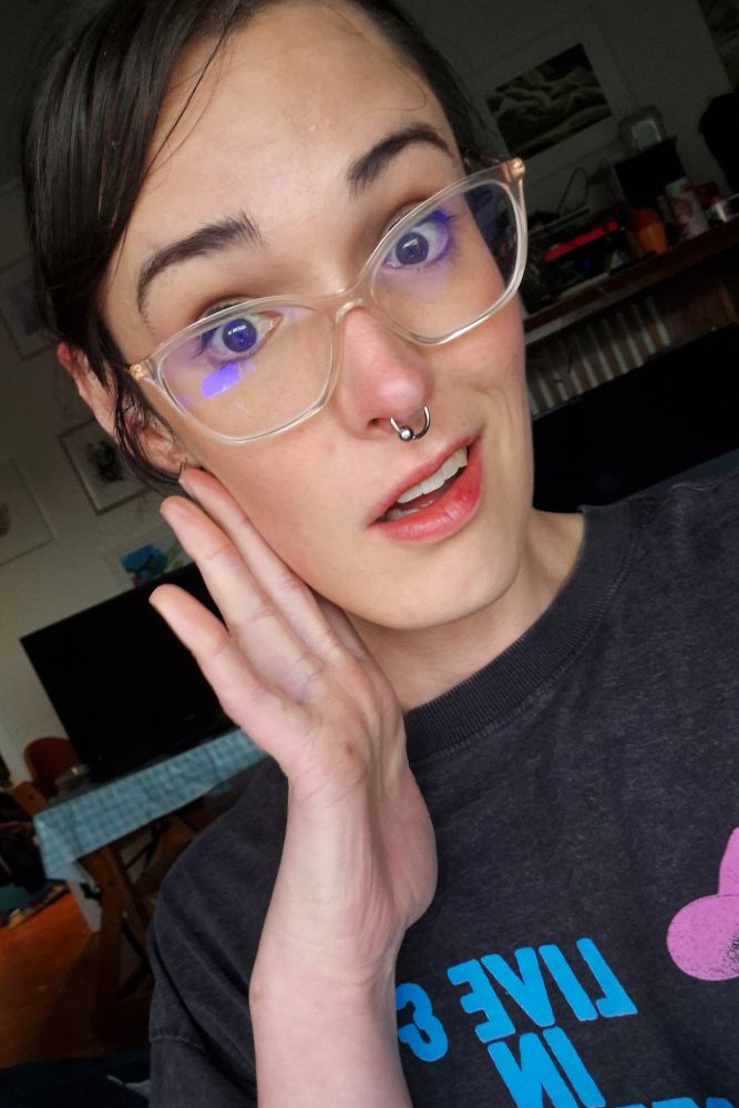 A photo of a woman wearing glasses, a dark grey shirt and makeup trying her best to look like she has two brain cells to rub together 