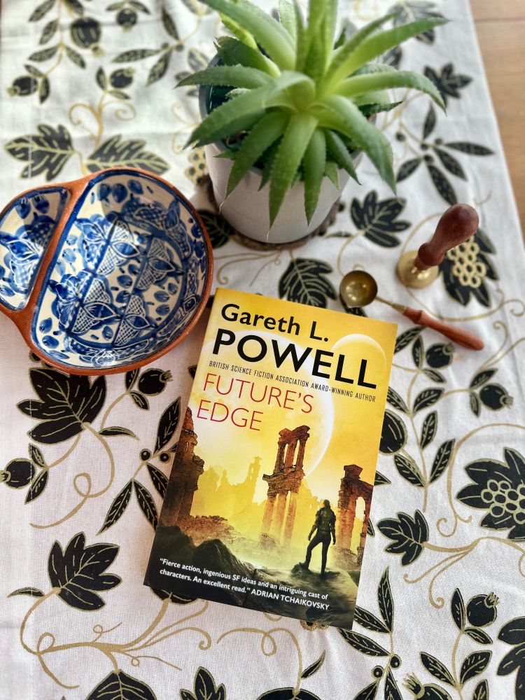 A copy of Future's Edge by Gareth L Powell on an embroidered table runner with a house plant, a wax seal kit, and an olive dish