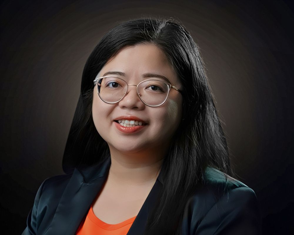 Ngan Tran has long black hair, wire-rimmed glasses, and is wearing a black suit jacket over an orange shirt.