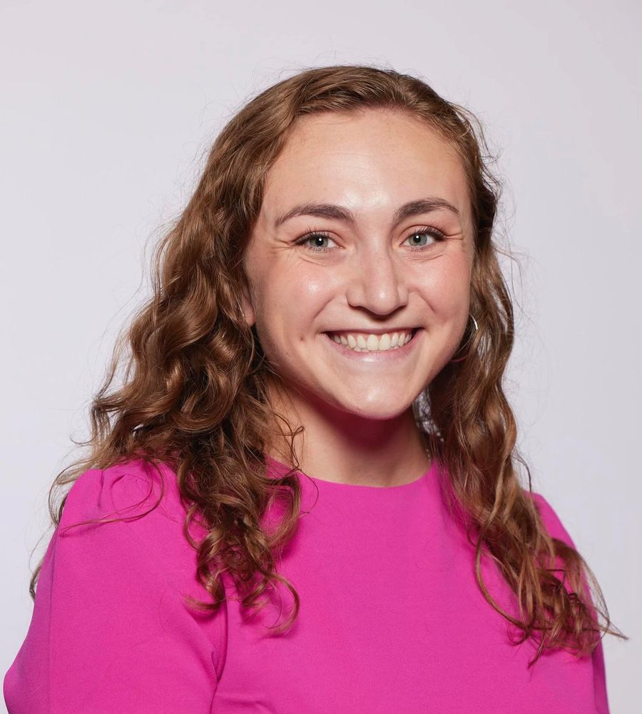 Molly Taylor has long curly brown hair and is wearing a bright pink shirt.