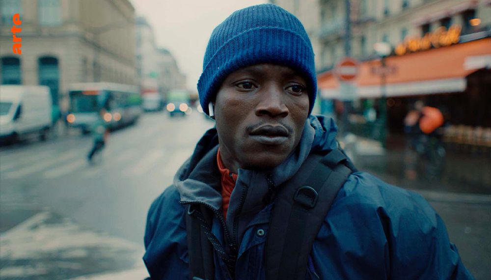 Photo d'illustration du film « L'histoire de Souleymane » sur laquelle on voit le comédien Abou Sangaré, il nous fait face, il porte une veste et un bonnet et se tient au milieu d'une rue, devant la Gare du Nord, derrière lui un bus, des passants et la devanture d'un restaurant.
