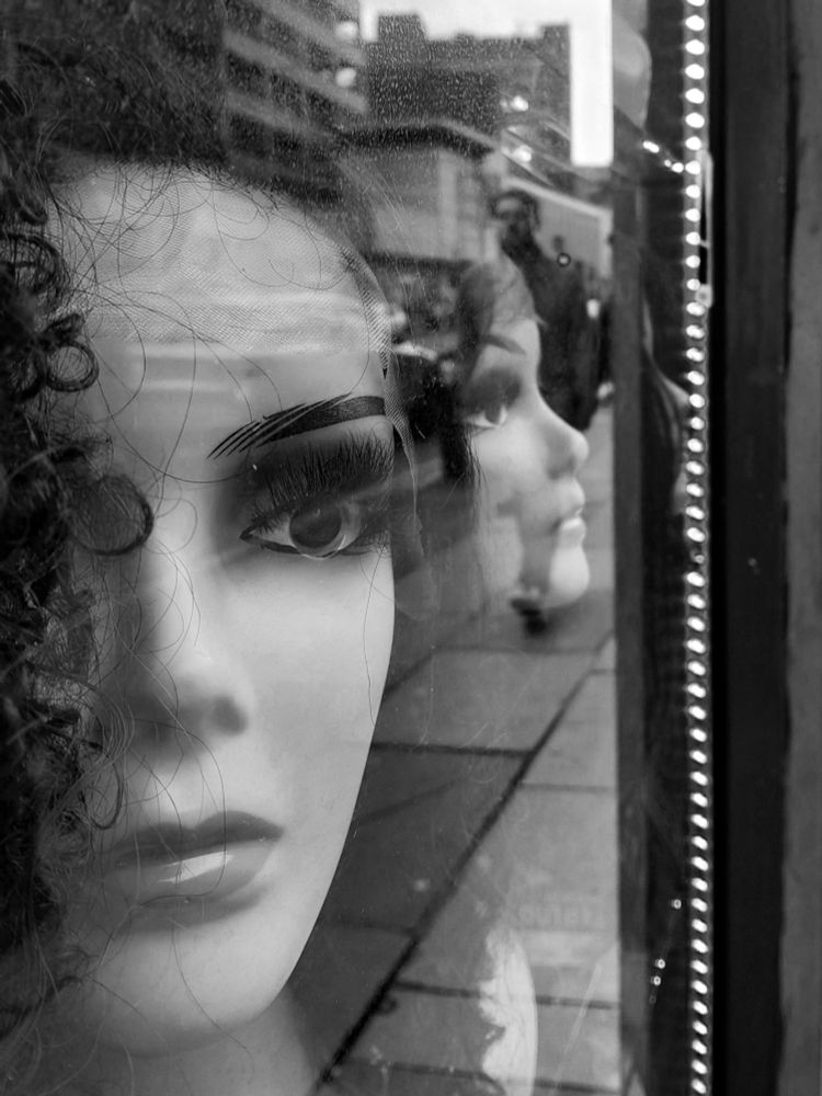 A photo looking into a hairdressers shop.
The window has mannequin heads modelling various wigs.
Two of the mannequins are looking out towards the street. 
A man walks past and is reflected in the window
A black and white photo 



