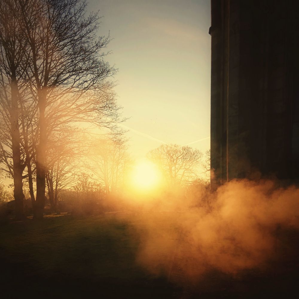 A photo of a hazy morning sunrise taken in a churchyard last week.
Bare wintery trees in a clear sky with the roll church on the right. 
Steam coming from a boiler looking like morning mist obscuring the golden sun in the distance 