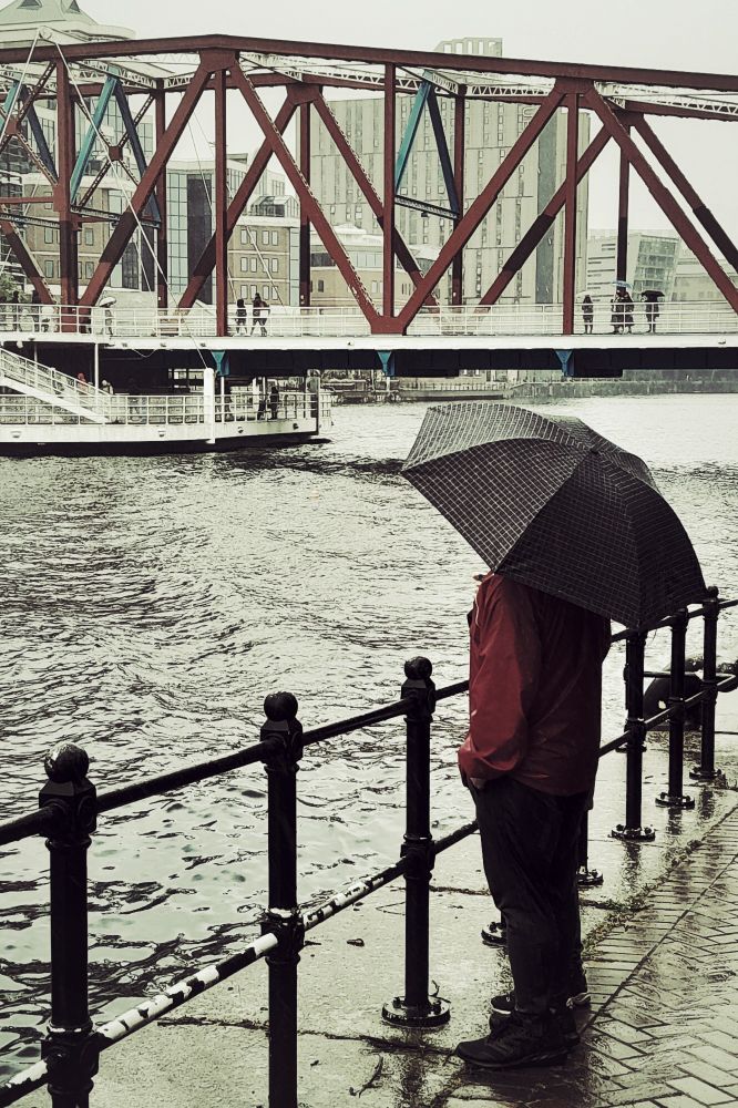 A man wearing a red jacket stands next to a metal fence at the Eerie Basin at Salford Quays. 
It is a rainy day and he is holding a black umbrella over his head.
In the distance is Big Red aka The Detroit Bridge that crosses the water.
People can be seen walking across the bridge 