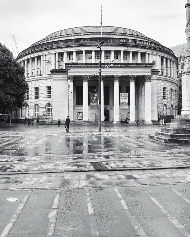 A black and white photo of Manchester Central Library. 
Looking across St Peter's Square towards the imposing circular library.
At the front are six romanesque pillars at the entrance.
Thd bottom level has arched windows and the second level has rectangular ones.
The top floor has pillars all around and there is a slightly domed roof.
It was a rainy day with the pavement glistening. 
The square is quiet with a man walking towards the library wearing all black with a trilby hat and briefcase


